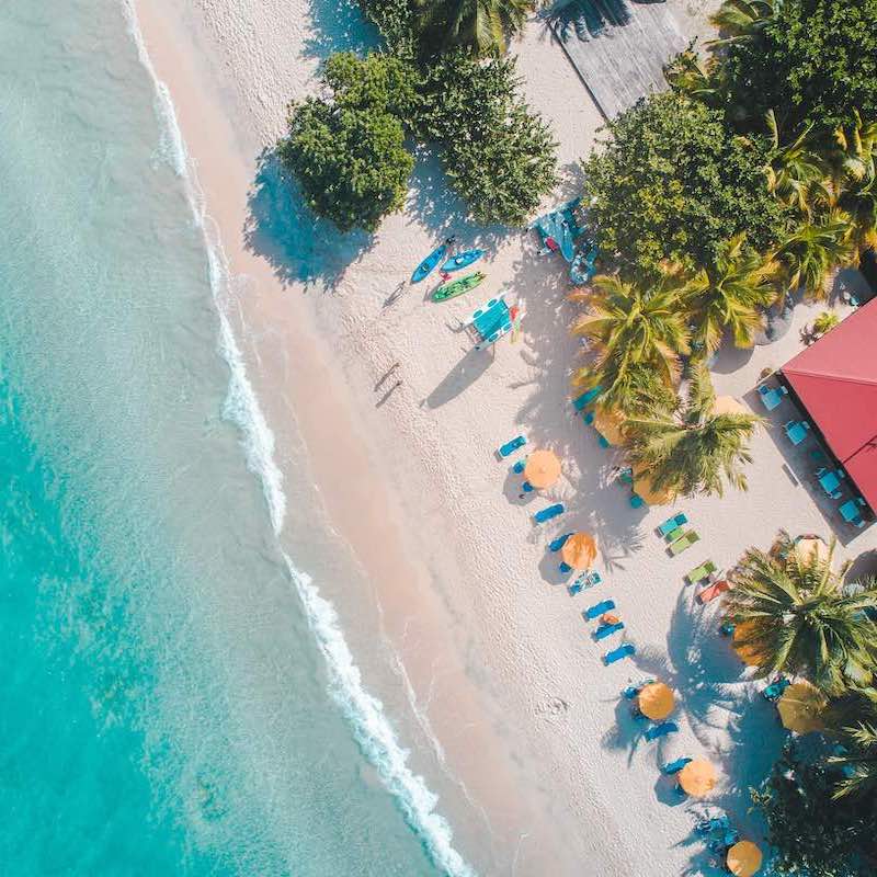 Imagen aérea de una de las playas para nómadas digitales en donde se aprecia la orilla, palmeras, sillas y toallas y el agua cristalina del mar