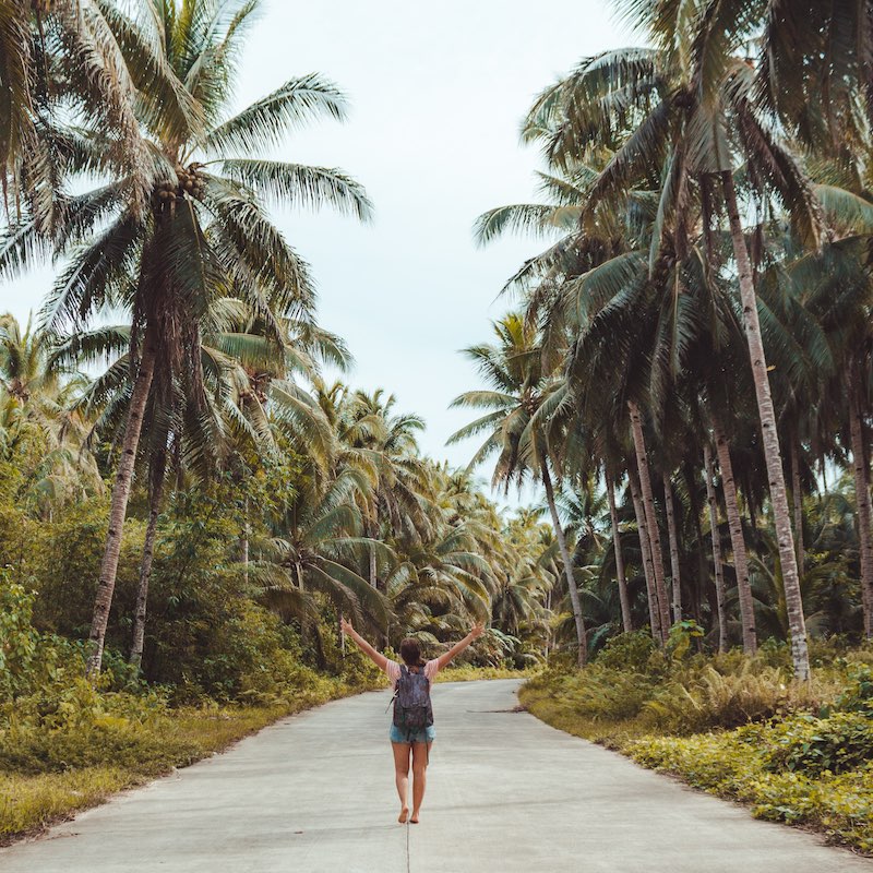 Mujer descalza caminando sobre carretera de asfalto en una ruta rodeada de palmeras con los brazos extendidos y una mochila, de espalda a la cámara