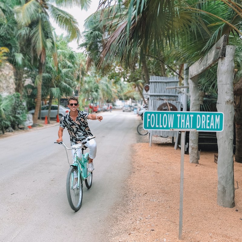Hombre con playera tropical en bicicleta señalando un cartel que dice "Follow that dream"