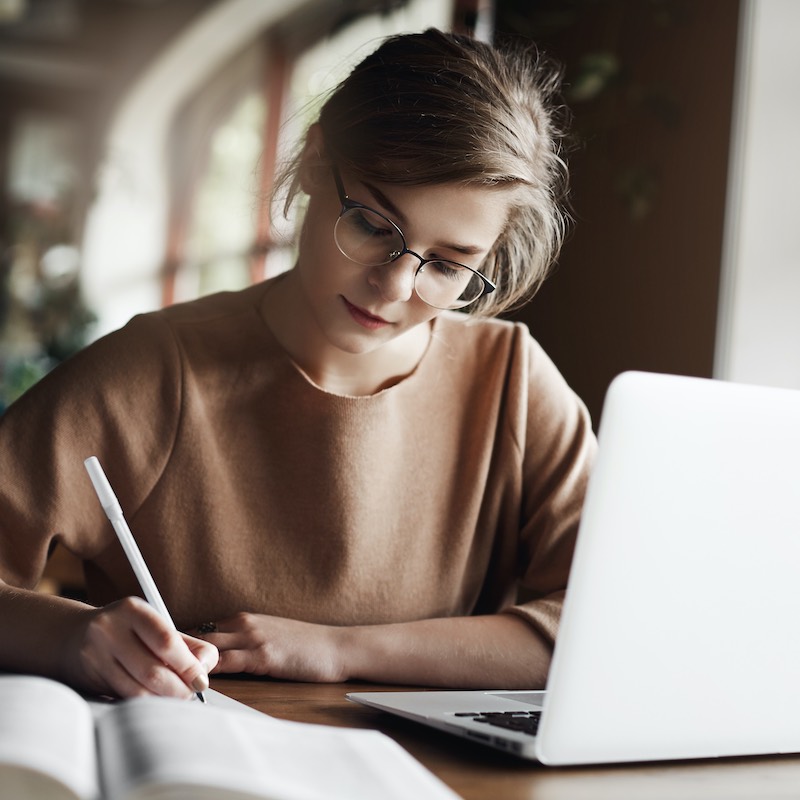 mujer concentrada escribiendo en papel y trabajando con su computador