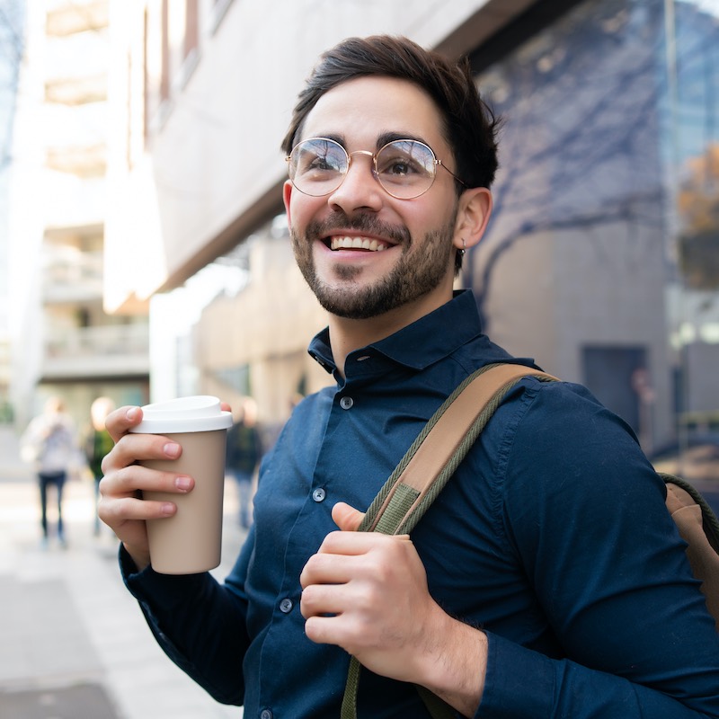 Joven sonriente con taza de café y mochila en la calle. Algunos ingenieros, consultores y desarrolladores web son de los que mejores ingresos alcanzan.