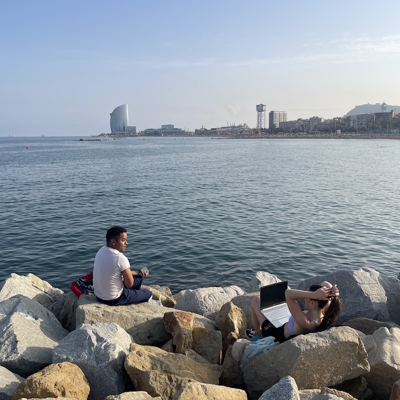 mujer con ropa deportiva trabajando con computadora unas sobre rocas mientras conversa con un hombre en Barcelona y admira el mar y el hotel W en la Barceloneta.jpg