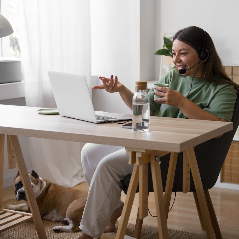 Mujer sentada en mesa de madera con su computadora en una videollamada junto a su perro