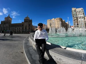 Fotografía de un hombre con camisa blanca y gafas de sol sentado junto a una fuente con su laptop sobre las piernas en una ciudad española