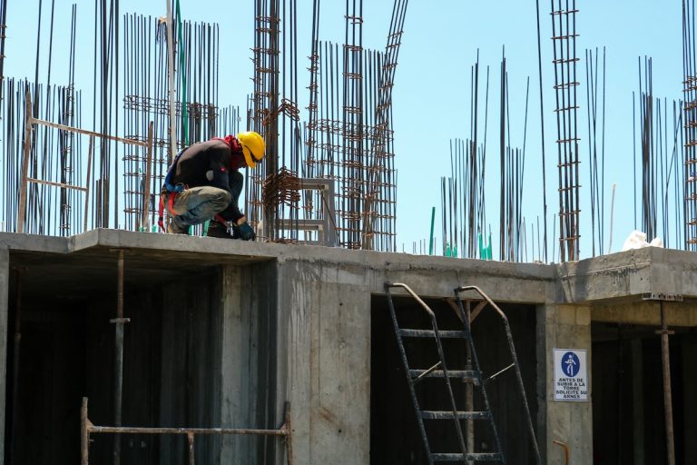 Obrero con casco amarillo trabajando agachado en lo alto de un edificio en construcción, rodeado de varillas de acero expuestas